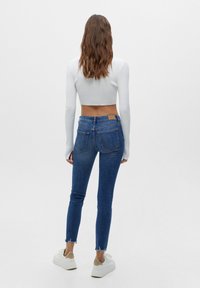 Jeans de mezclilla azul ajustados con dobladillos desflecados en los tobillos, combinados con una blusa blanca de manga larga recortada, llevada con zapatillas blancas.