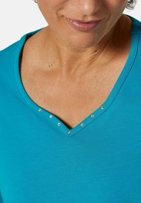 Turquoise V-hals top van zachte stof, met decoratieve zilveren studs langs de halslijn en een gladde textuur.