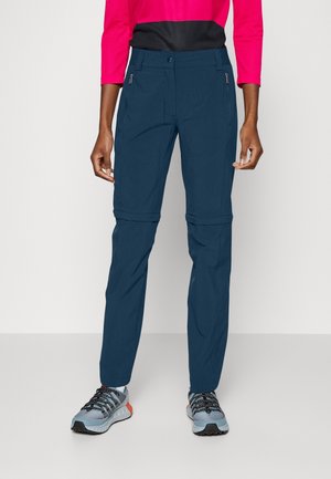 Person trägt marineblaue Reißverschluss-Hose mit abnehmbaren unteren Beinen, ein rot-schwarzes Langarmshirt und graue Wanderschuhe mit schwarzen Schnürsenkeln.