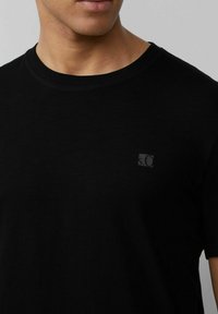 Schwarzes Baumwoll-T-Shirt mit rundem Halsausschnitt und kurzen Ärmeln, ausgestattet mit einem kleinen, passenden Logo-Patch auf der linken Seite der Brust.