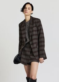 Brauner karierten Blazer mit grauen Streifen, kombiniert mit einem strukturierten grauen Pullover und einem passenden kurzen Rock, ergänzt durch kniehohe schwarze Stiefel.