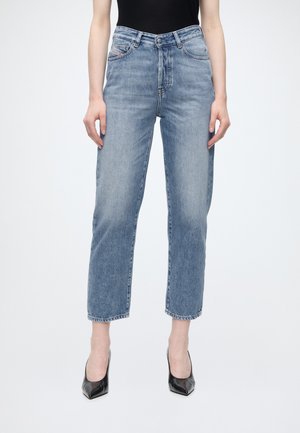 Femme portant un jean bleu taille haute à jambes droites et des talons noirs à bouts pointus, debout sur un fond blanc.