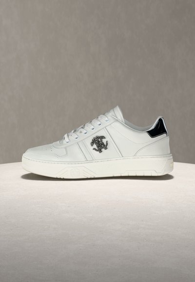 Λευκό χαμηλό αθλητικό παπούτσι με μαύρη πινελιά στη φτέρνα, κορδόνια στο μπροστινό μέρος και μαύρο κεντημένο λογότυπο στο πλάι, τοποθετημένο πάνω σε ανοιχτή υφή επιφάνειας.