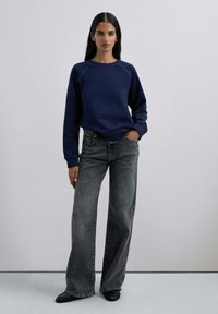 Marineblaue Sweatshirt mit Raglanärmeln, kombiniert mit hoch taillierten, weit geschnittenen, ausgewaschenen schwarzen Jeans und schwarzen Schuhen. Glatte Textur, minimales Design.