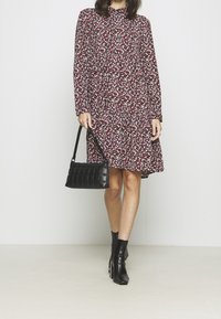 Červené šaty s květinovým vzorem, dlouhými rukávy a volánkovým spodním lemem, doplněné černou prošívanou kabelkou a černými kotníkovými botami.