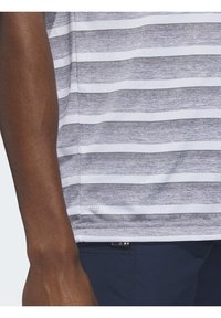 Detailný záber na ruku osoby vedľa bielej a sivej prúžkovanej košele a tmavomodrých nohaviciach s mal�ým zipsom.