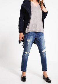 Femme portant un manteau bleu marine, un pull à col en V gris clair, un jean bleu déchiré et des chaussures noires sans lacets, debout devant un fond blanc.