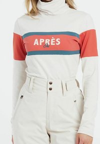 Frau trägt einen weißen Rollkragenpullover mit rot-blauen Streifen und dem Text "Après Ski", kombiniert mit hoch-taillierten weißen Schneehosen mit Reißverschlusstaschen.