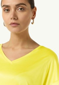 Żółta koszulka z dekoltem w serek, wykonana z miękkiego materiału, o luźnym kroju. Modelka nosi minimalne dodatki, z dużymi złotymi kolczykami w kształcie kół.