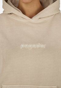 Beżowa bluza z kapturem z przednią kieszenią i haftowanym logo "pegador" w dopasowanym kolorze na klatce piersiowej. Miękka bawełniana struktura.