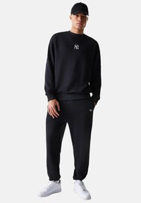 Felpa nera con logo bianco, abbinata a pantaloni jogger neri e scarpe da ginnastica bianche. Il tessuto sembra morbido con una vestibilità comoda.