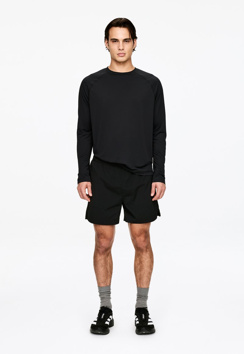 Zwart atletisch shirt met lange mouwen gecombineerd met zwarte korte broek, grijze sokken en zwarte sneakers met witte strepen. Casual sportoutfit.