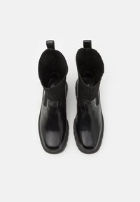 Botine negre cu un upper din piele lucioasă, guler din tricot cu fante, și talpă din cauciuc. Au bucle pentru a fi îmbrăcate și dezbrăcate cu ușurință.