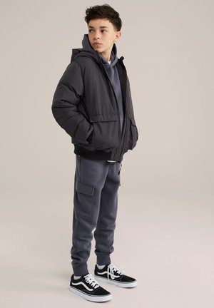 Giacca a vento nera con cappuccio, felpa grigia sotto, e pantaloni della tuta grigio scuro con tasche. Sneakers bianche e nere completano l'outfit.