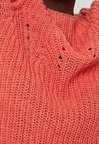 Korale sweter z dzianiny o luźnym, teksturowanym splocie z małymi otworami, podkreślający swoją miękką tkaninę i swobodny krój.