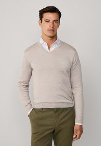 Młody mężczyzna w beżowym swetrze z dekoltem w serek na białej koszuli z kołnierzykiem i oliwkowych spodniach, stoi z jedną ręką w kieszeni.