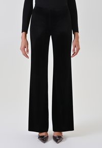 Pantaloni ampi in velluto nero con una texture liscia, vita alta e lunghezza fino al pavimento. Abbinati a décolleté a punta con fiocco.