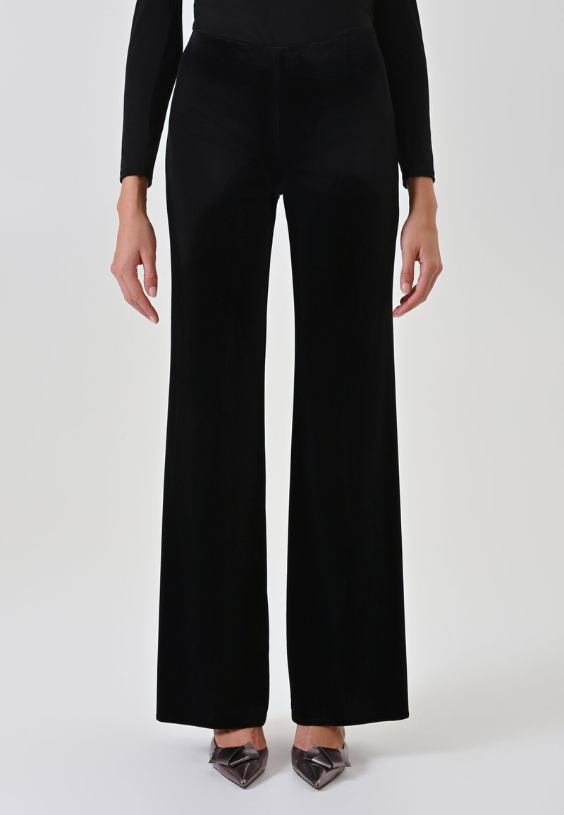 Pantaloni ampi in velluto nero con una texture liscia, vita alta e lunghezza fino al pavimento. Abbinati a décolleté a punta con fiocco.