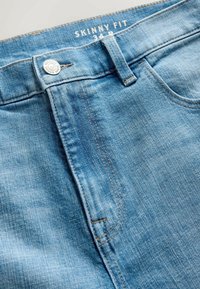 Blaue Skinny-Jeans mit Metallknopf und sichtbarer Gürtelschlaufe, Größenetikett 34 R innen am Bund.