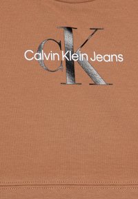 Ruda medvilnė T-marškiniai su išryškintu juodu ir sidabriniu "CK" logotipu ir tekstu "Calvin Klein Jeans" apačioje. Lygus paviršius, apvalus kerinys.