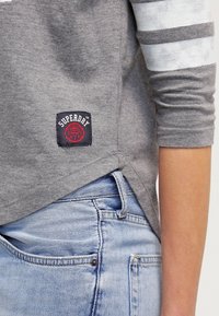 Grå bomulls långärmad skjorta med en tagg som visar "Superdry"-logotypen, i kombination med ljusblå denimjeans som har en framficka och sömnadsdetaljer.