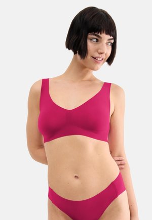 Femme aux cheveux courts et foncés portant un ensemble soutien-gorge et culotte rose assorti, regardant sur le côté avec un léger sourire.
