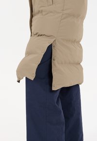 Beige pufferjakke med trykknapper i siden, parret med mørkeblå bukser. Jakken har en glat tekstur og en let oversized pasform.