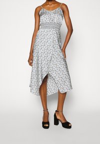 Femme portant une robe sans manches, longue jusqu'aux chevilles, à motifs, cintrée à la taille, avec des sandales noires à plateforme à bout ouvert et à brides à la cheville.