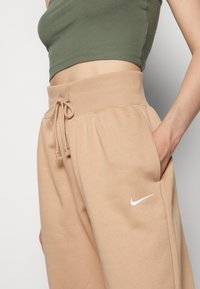 Béžové tepláky s rebrovaným pásom a šnúrkou na stiahnutie. Obsahujú bočné vrecká a malé biele logo Nike na ľavom stehne.