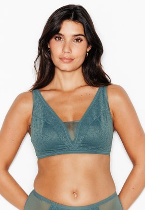 Femme aux longs cheveux foncés portant un bralette en dentelle bleu sarcelle et une lingerie assortie, debout devant un fond blanc, faisant face à l’avant.
