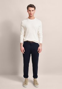 Cremfarbener Pullover, navyfarbene Hose, beige Sneaker. Der Pullover hat einen Rundhalsausschnitt. Die Hose hat einen lässigen Schnitt mit umgeschlagenen Säumen.
