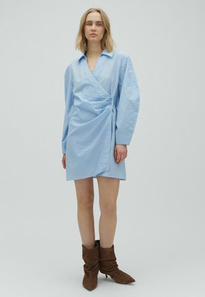 Kvinde med skulderlangt blondt hår iført en lyseblå slå-om-kjole med lange ærmer og brune ruskindsstøvler, der står foran en ensfarvet baggrund.