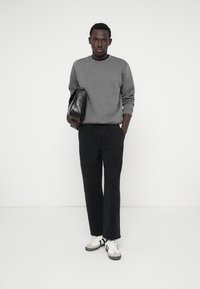 Sivá mikina, čierne voľné nohavice a biele tenisky s čiernymi akcentmi; drží malú čiernu tašku. Jednoduchý dizajn, neutrálné farby.