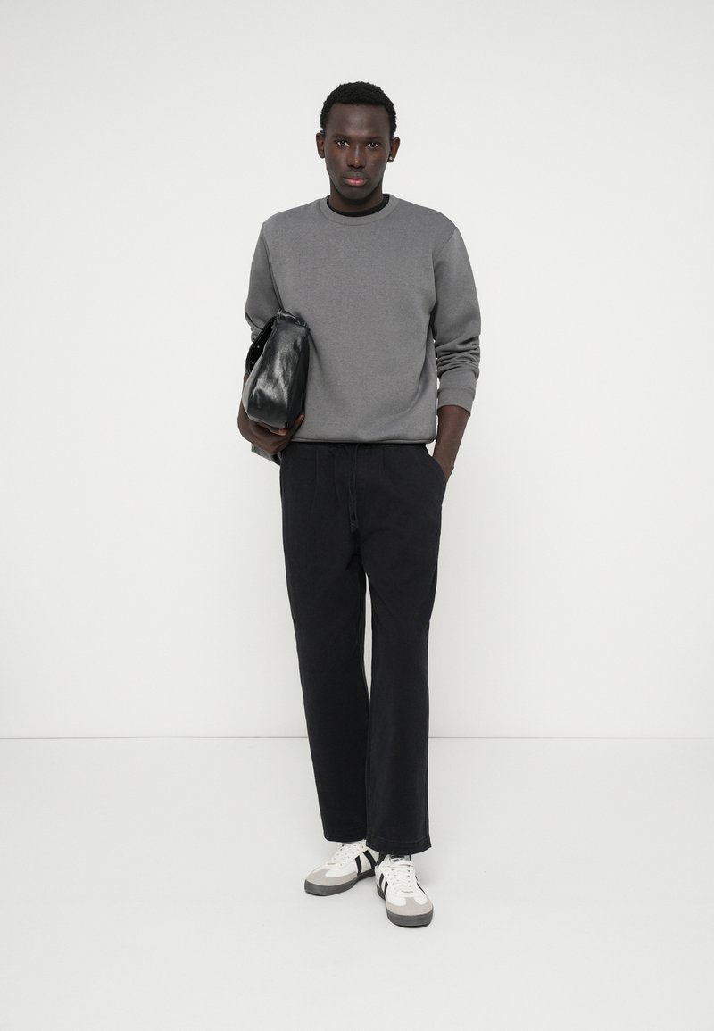 Sivá mikina, čierne voľné nohavice a biele tenisky s čiernymi akcentmi; drží malú čiernu tašku. Jednoduchý dizajn, neutrálné farby.