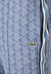 Blå mönstrade joggers i texturerad tyg, med en geometrisk design och kontrasterande sidostripe. Inkluderar en liten grön krokodil-logotyp.