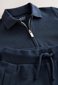 Polo bleu marine zippé avec col et poignets côtelés ; doté d'une fermeture éclair argentée et d'un tissu texturé, associé à un pantalon de survêtement à cordon de serrage assorti.