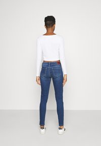 Jeans ajustados de color azul con una cintura a media altura, que presentan bolsillos traseros y detalles de costura sutiles. Combinados con una blusa de manga larga blanca.