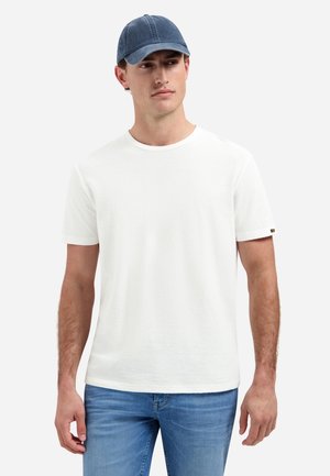 Mann trägt ein schlichtes weißes T-Shirt mit kurzen Ärmeln, blaue Jeans und eine dunkelblaue Baseballkappe, steht mit neutralem Gesichtsausdruck vor weißem Hintergrund.