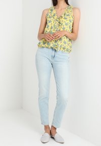 Gul blommig ärmlös topp med volangdetalj, i kombination med ljusblå slim fit jeans och grå slip-on skor.