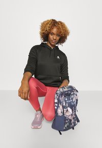 Virágmintás hátizsák kék és rózsaszín kiegészítőkkel, fekete kapucnis pulóverrel és rózsaszín leggings-szel viselve. A sportcipő könnyű lila mintázattal rendelkezik.
