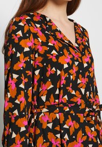 Kvinna i en svart klänning med orange och rosa blommönster, V-ringad krage och ett knytbälte i midjan.