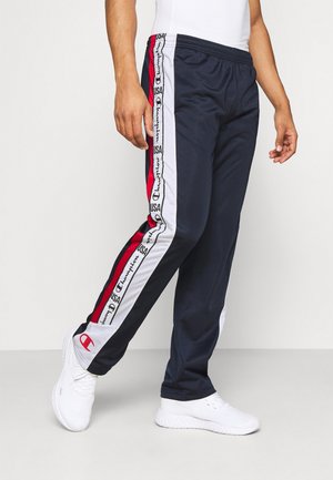 Mann trägt marineblaue Champion-Trainingshose mit roten und weißen Streifen und Logos, kombiniert mit weißen Sportschuhen.