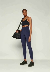 Mujer con sujetador deportivo negro, leggings de tiro alto azul marino y zapatillas negras, sosteniendo una bolsa de gimnasio negra sobre su hombro frente a un fondo liso.