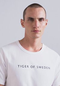 Ung mann med kort hår iført en hvit t-skjorte med teksten "TIGER OF SWEDEN", stående mot en ensfarget lys grå bakgrunn.