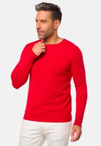 Homme aux cheveux courts et foncés, barbu, portant un pull rouge à manches longues et un pantalon blanc, regardant vers la gauche, la main près du cou.