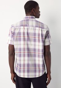 Mees, kes kannab lühikeste varrukatega lilla, valge ja punase ruudulise mustriga särki, vaadatuna tagant valge tausta ees.
