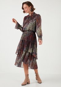 Langes, schichtweise, durchsichtiges Kleid in tiefem Kastanienbraun mit filigranen Paisleymustern in Grün, Orange und Blau. Mit einer gebundenen Halslinie und weiten Ärmeln.