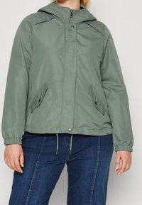 Light green lightweight jacket with a hood, zip closure, two side pockets, and elastic cuffs. Smooth texture and casual fit.
