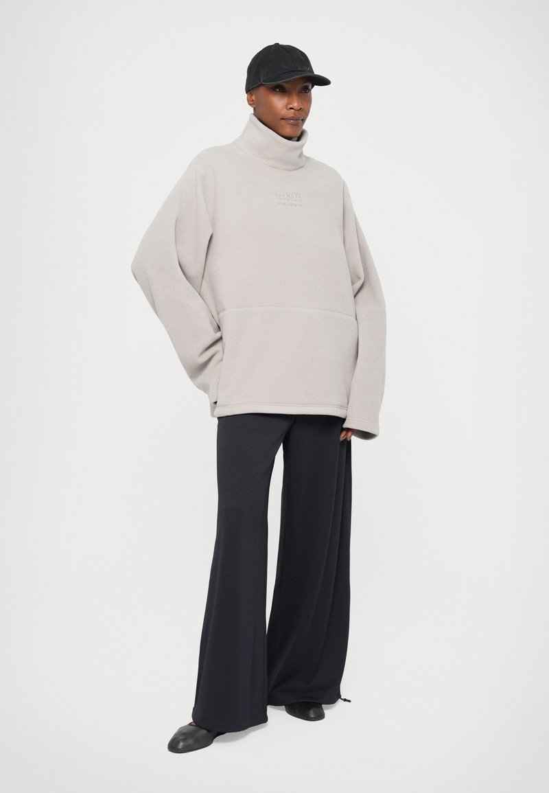 Persoon in een lichtgrijze oversized coltrui van fleece, brede zwarte broek, zwarte pet en zwarte schoenen, staand tegen een vlakke achtergrond.