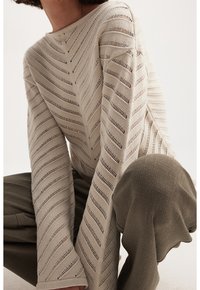 Beige neulepaita, jossa siksak-kuvio ja avoimet ompeleet. Malli yhdistää sen oliivinvihreisiin, teksturoiduihin housuihin, kyykkyasennossa.